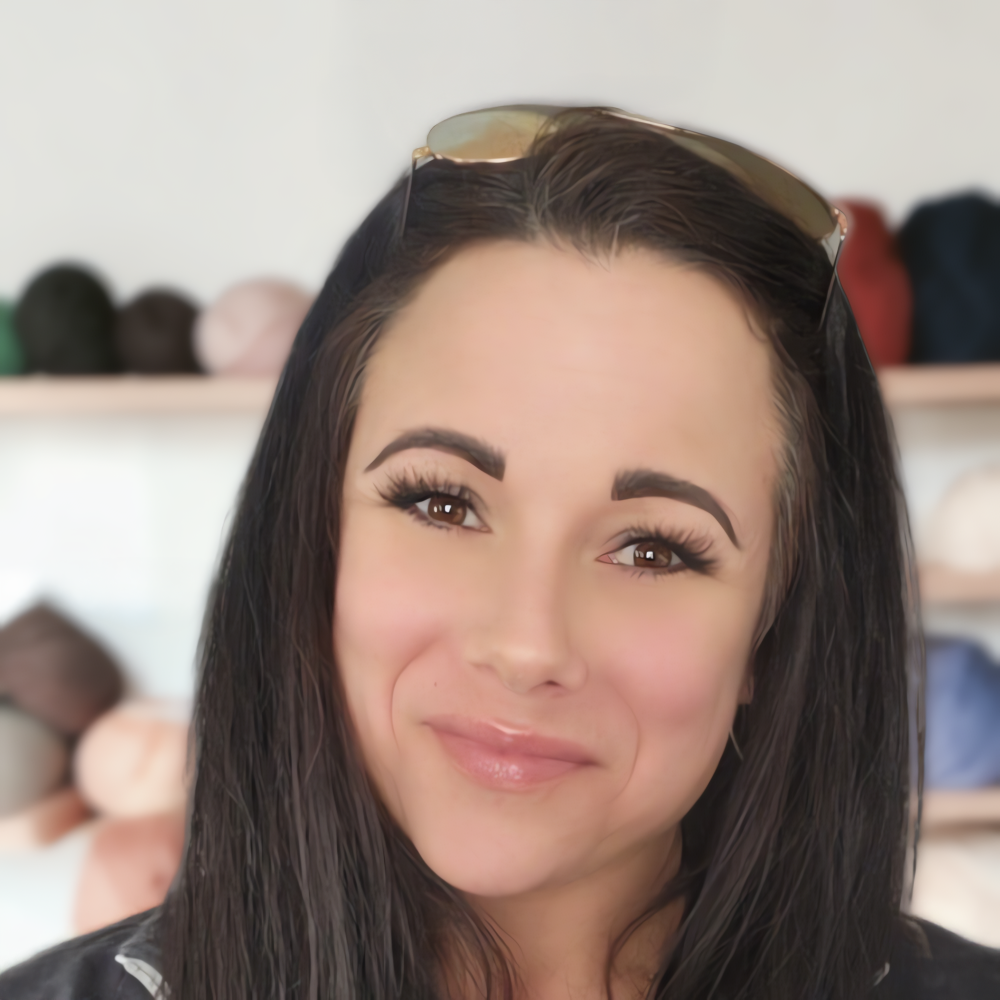 A close-up portrait of a woman with long dark hair wearing sunglasses on top of her head, set against a softly blurred background with shelves of colorful yarns.