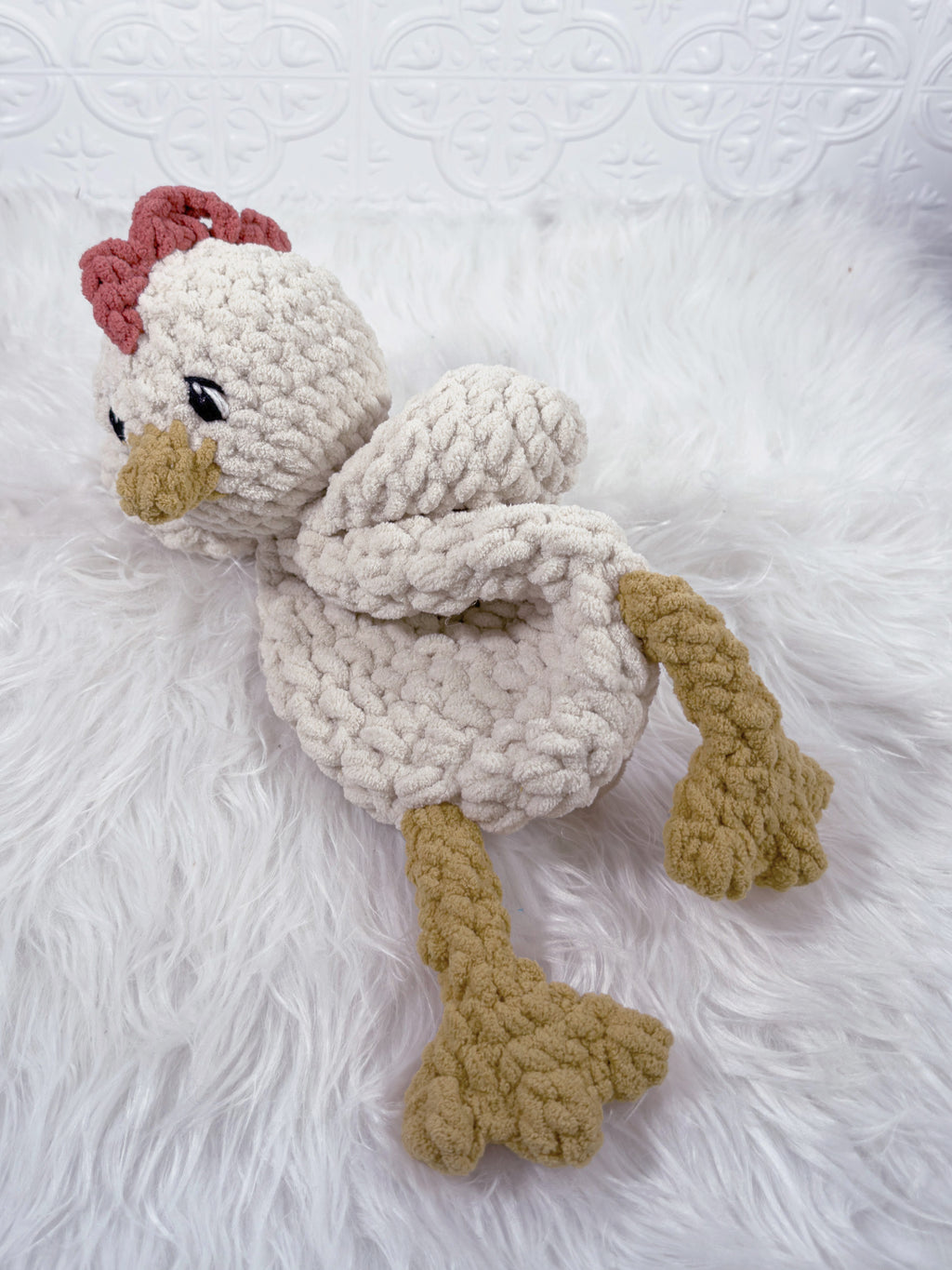 A crocheted plushie chicken snuggler security blanket textured off-white feathers, tan legs and beak, and a dusty red comb, resting on a white faux fur surface.