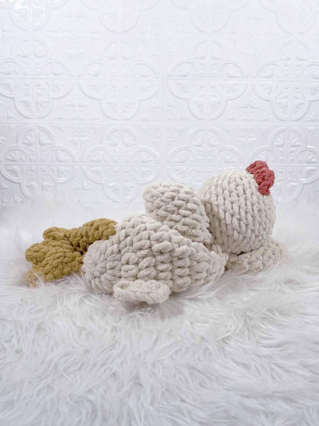 A handmade crocheted chicken snuggler security blanket lying on a white faux fur surface against a white patterned backdrop. The chicken is primarily off-white/cream with a mustard yellow comb and tail, and a small red comb accent.