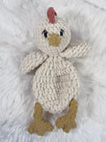 A crochet plushie chicken snuggler security blanket with a cream-colored body, tan beak and feet, and a small red comb, lying on a white faux fur background.