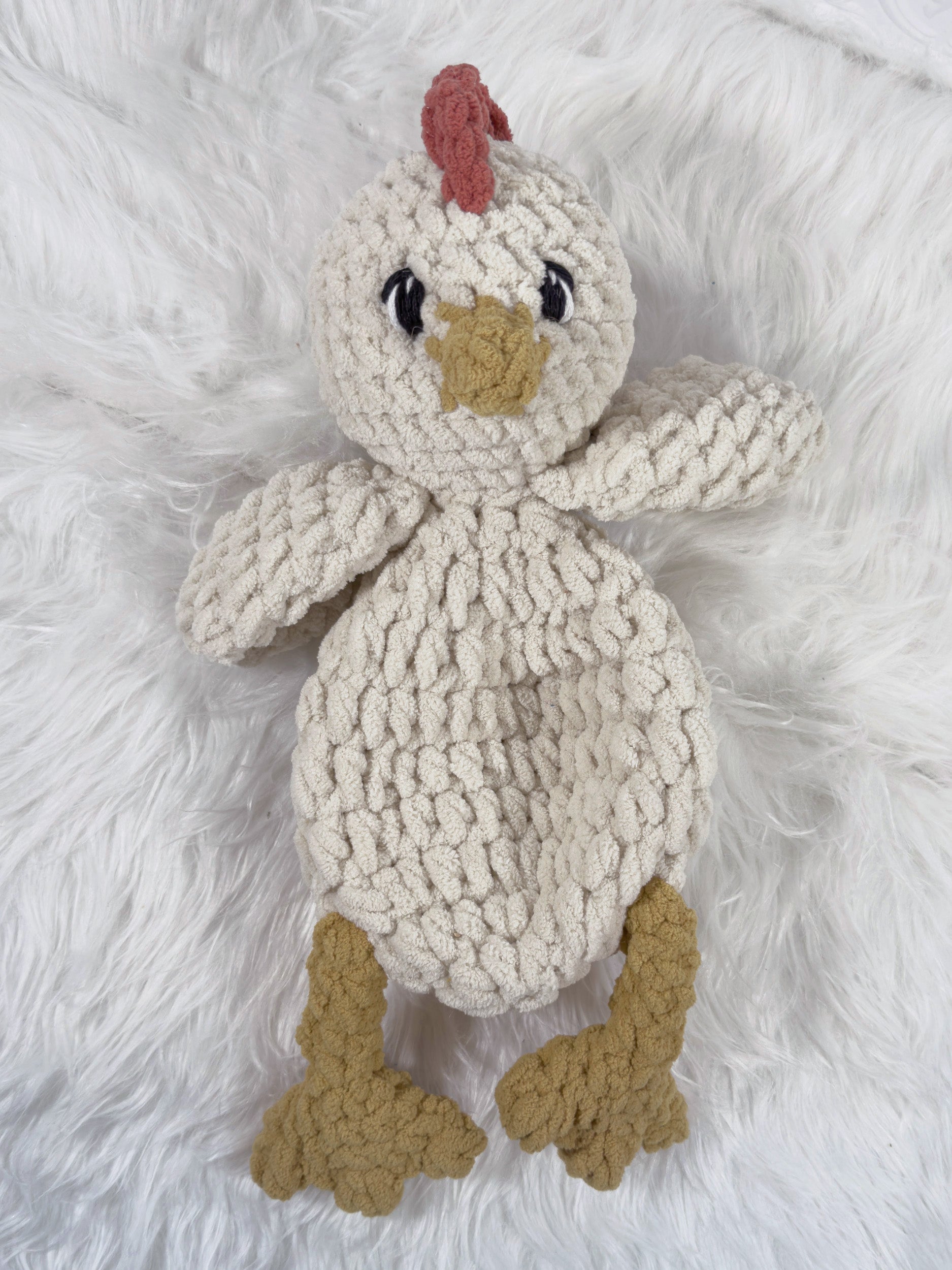 A crochet plushie chicken snuggler security blanket with a cream-colored body, tan beak and feet, and a small red comb, lying on a white faux fur background.