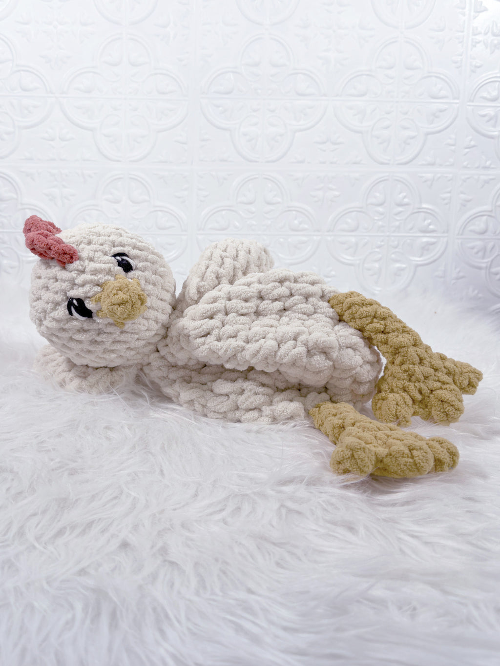 A handmade crocheted chicken security blanket or plush toy, featuring a chunky off-white body, yellow beak and feet, and a pink comb, resting on a white faux fur surface.
