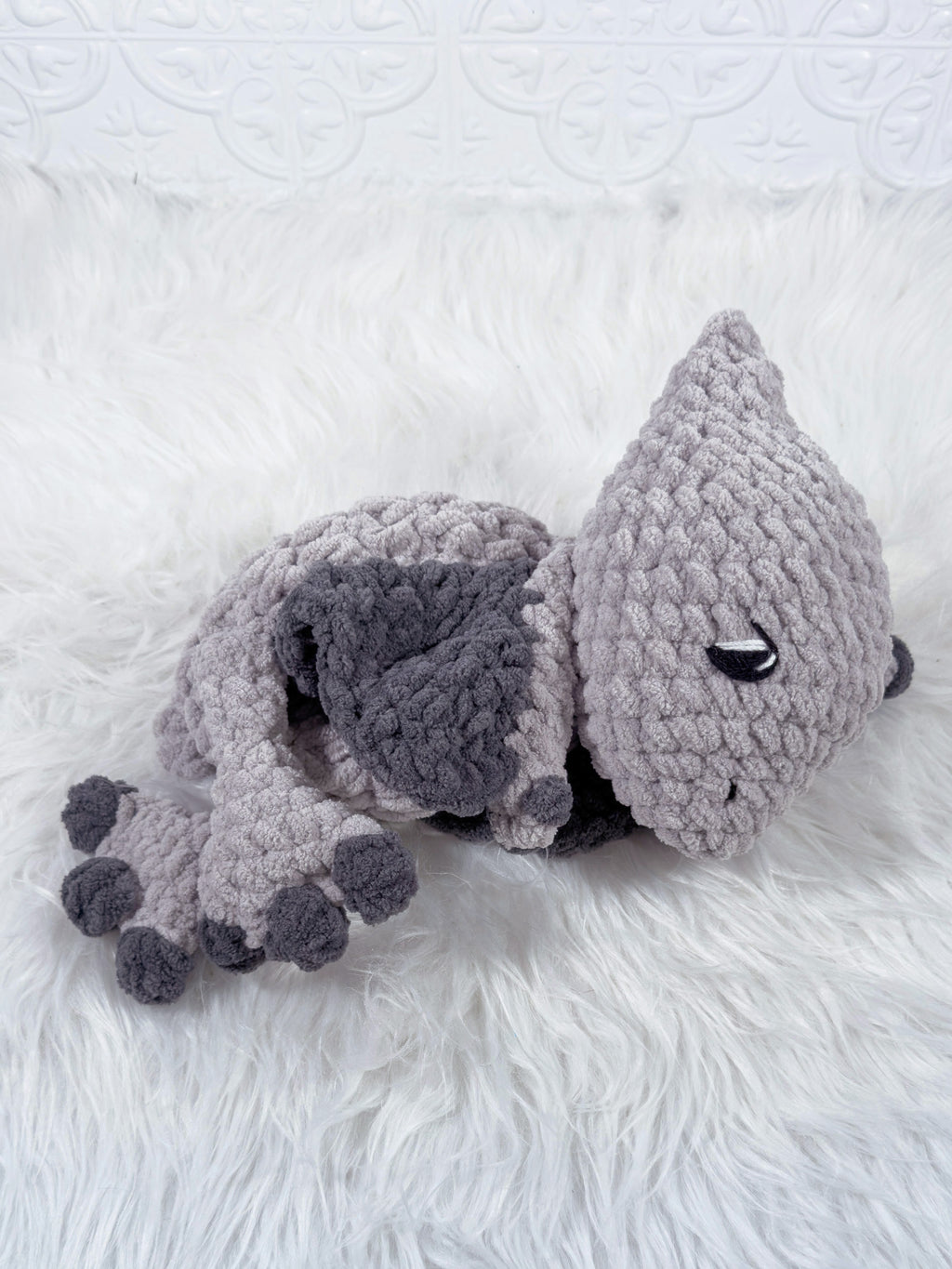 A handmade crocheted plush pterodactyl baby snuggler in light grey with dark charcoal accents, resting on a white faux fur surface.