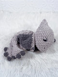 A handmade crocheted plush pterodactyl baby snuggler in light grey with dark charcoal accents, resting on a white faux fur surface.