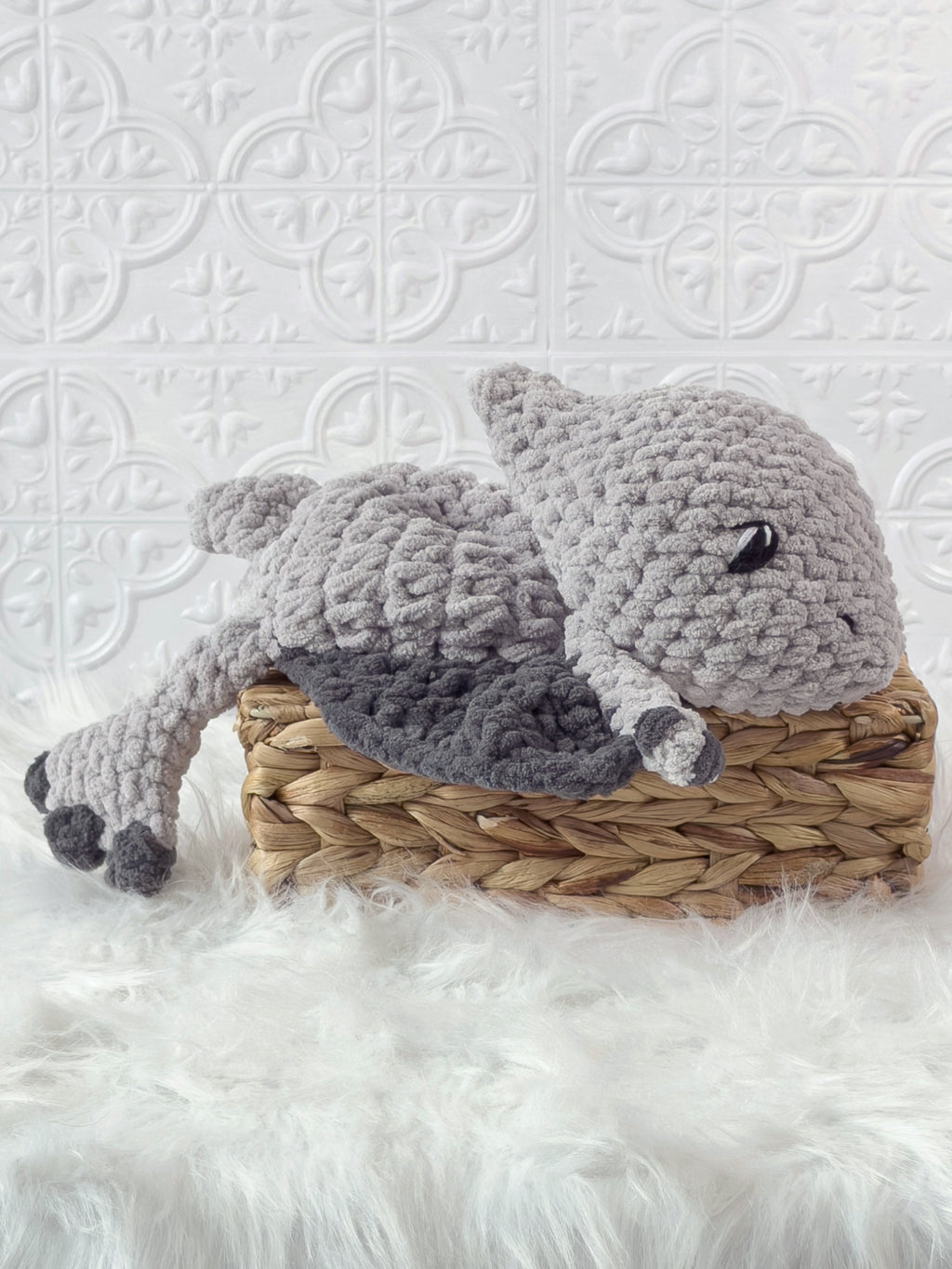 A crocheted plush animal toy, sleeping pterodactyl baby snuggler, resting on a woven wicker basket over a white faux-fur surface against a white embossed tile wall.