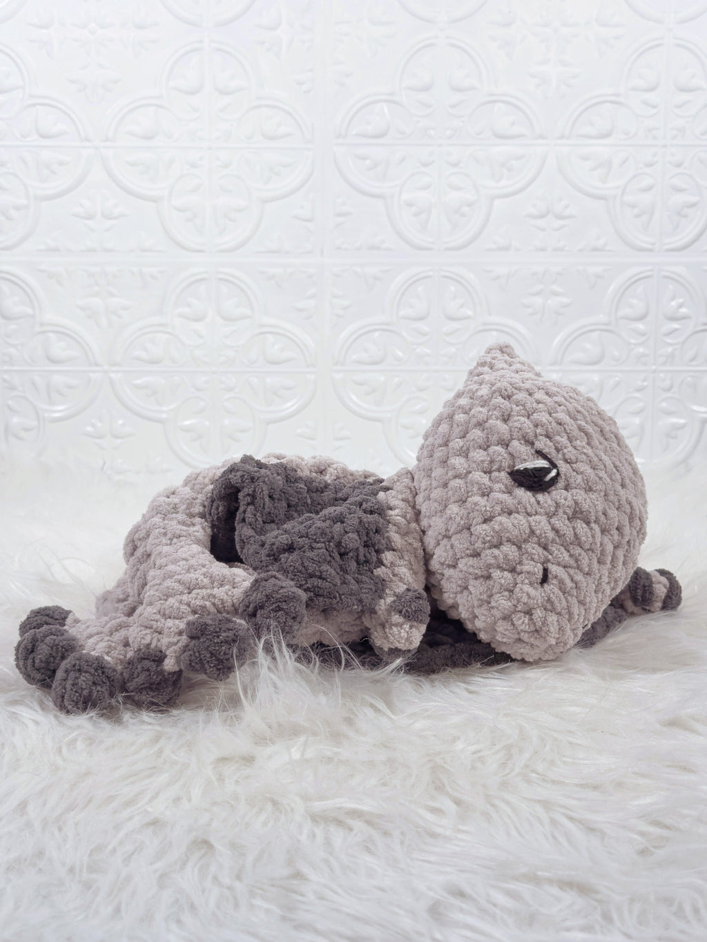 A handmade crocheted  pterodactyl baby snuggler plush toy in shades of light gray and dark gray, lying on a white faux fur rug against a white embossed tile wall.