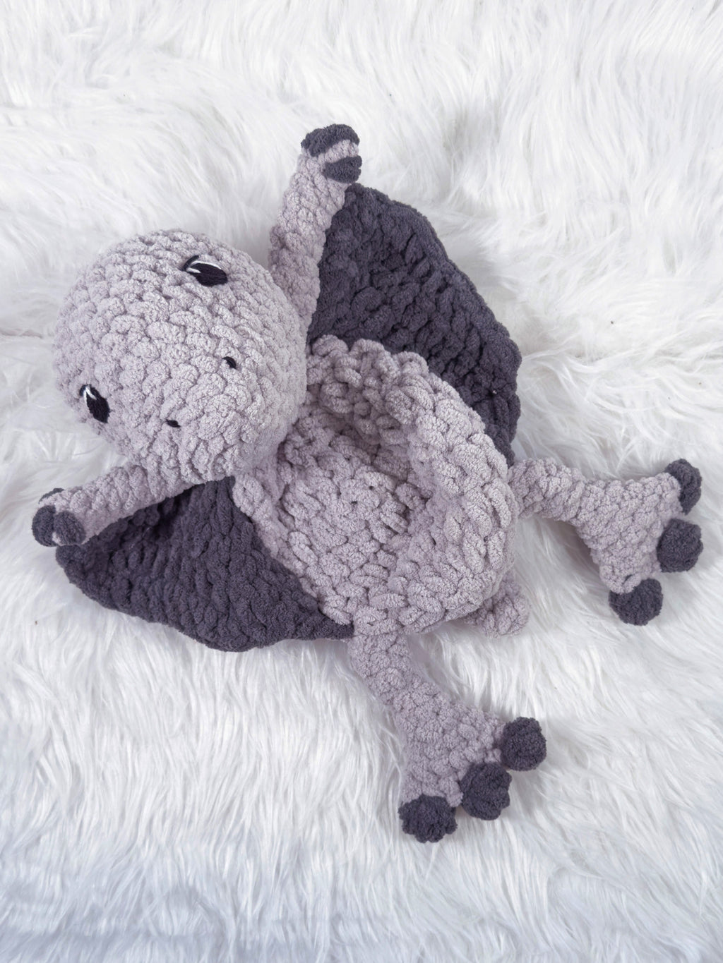 A handmade crocheted pterodactyl baby snuggler security blanket in light gray and dark gray, lying on a white faux fur surface.
