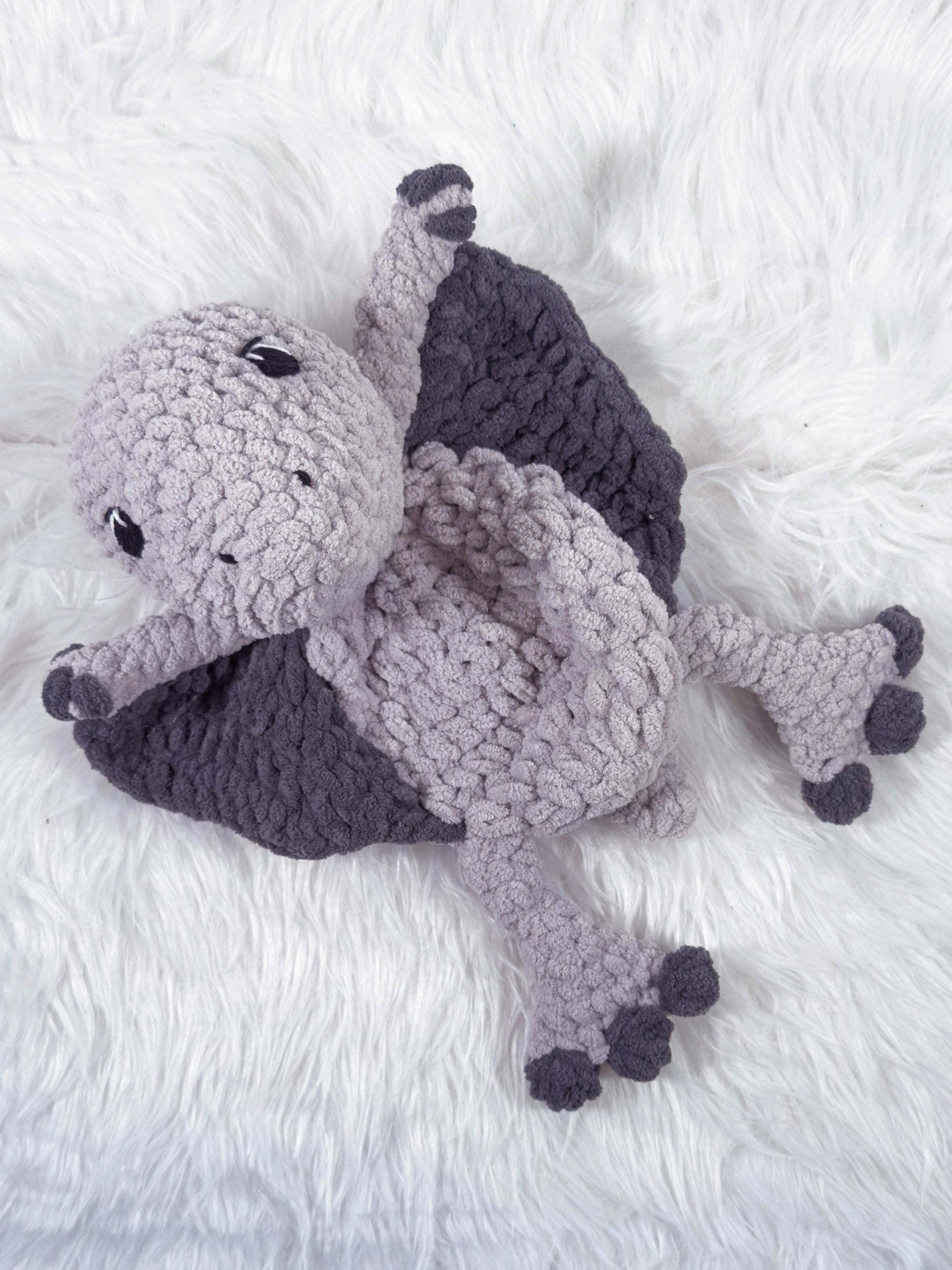 A handmade crocheted pterodactyl baby snuggler security blanket in light gray and dark gray, lying on a white faux fur surface.