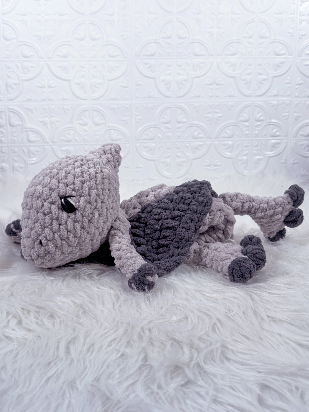 A handmade crocheted plush pterodactyl baby snuggler, lying on a white faux fur surface against a white embossed tile wall. The toy is primarily light gray with a darker gray back and black accents.