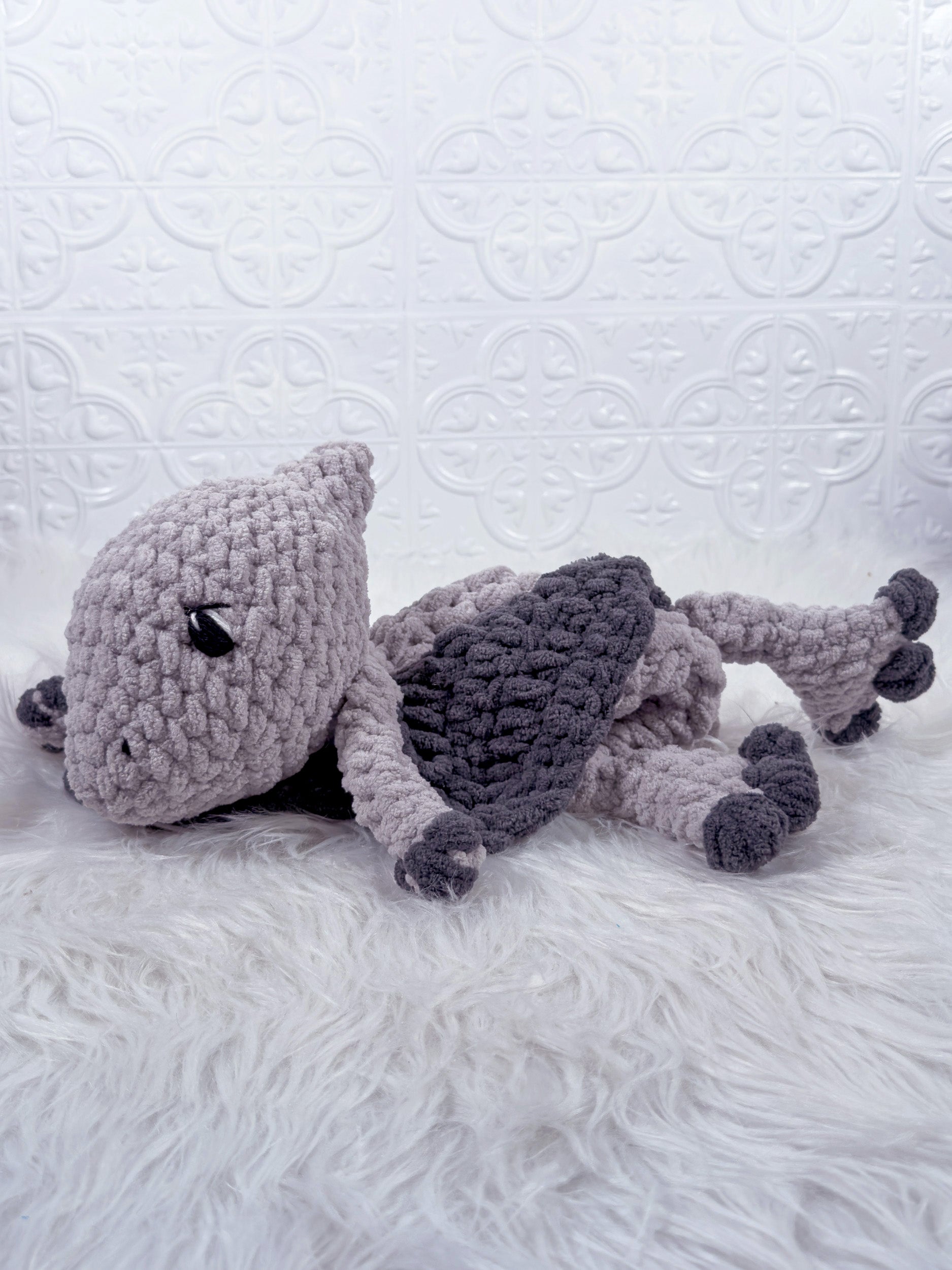 A handmade crocheted plush pterodactyl baby snuggler, lying on a white faux fur surface against a white embossed tile wall. The toy is primarily light gray with a darker gray back and black accents.