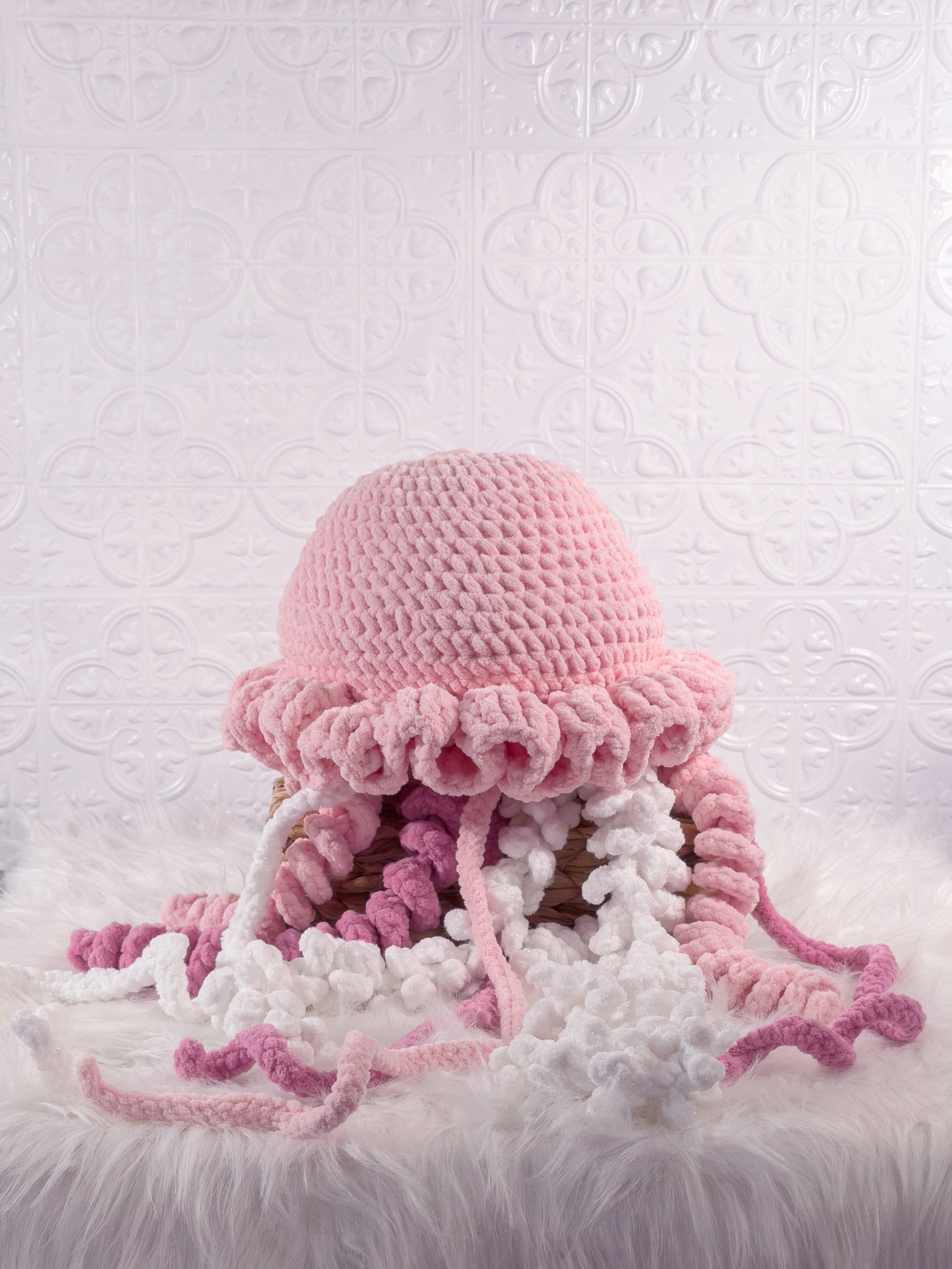 A pink and white crocheted jellyfish plushie toy with long tentacles, displayed on a white faux fur surface against a white embossed patterned backdrop.
