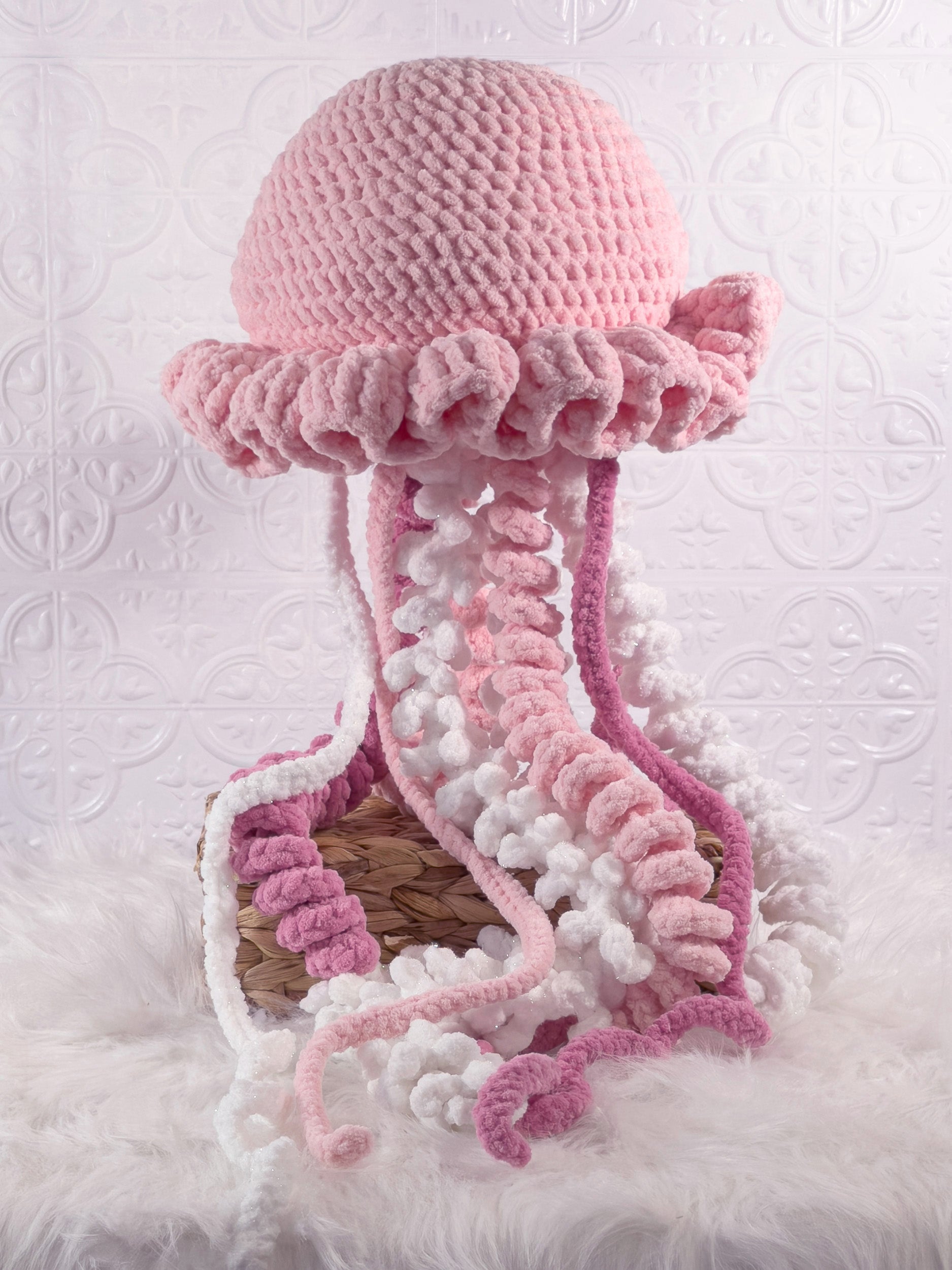 A crocheted jellyfish plushie featuring a pink textured dome and ruffled edge with long, wavy tentacles in pink, white, and light pink yarn, displayed on a white faux fur surface.
