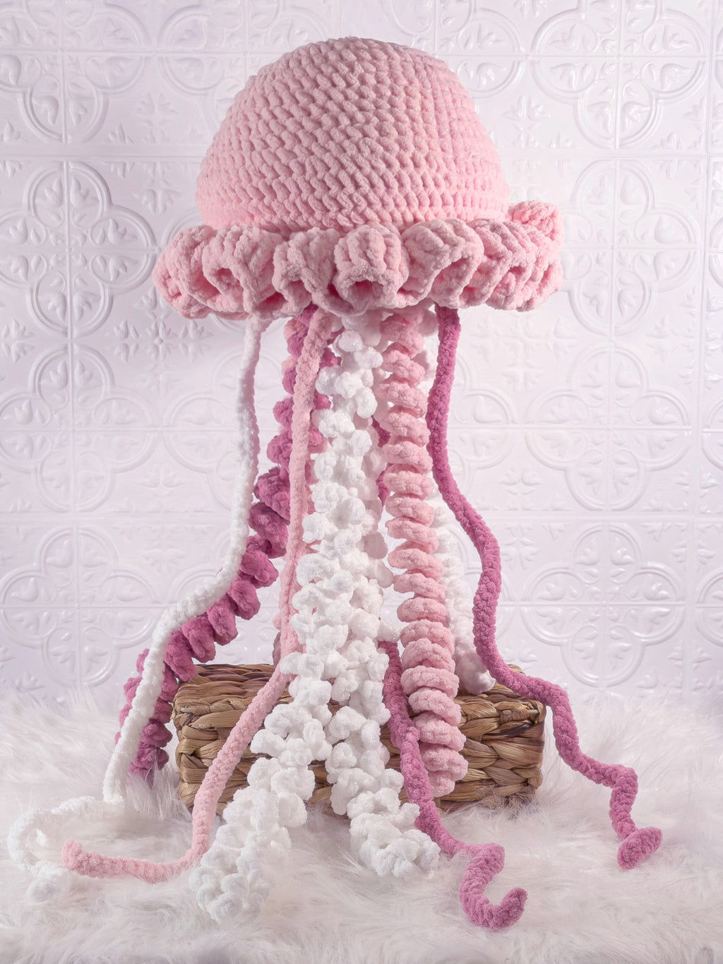 A crocheted jellyfish hat in shades of pink and white, displayed on a small woven basket against a textured white background.