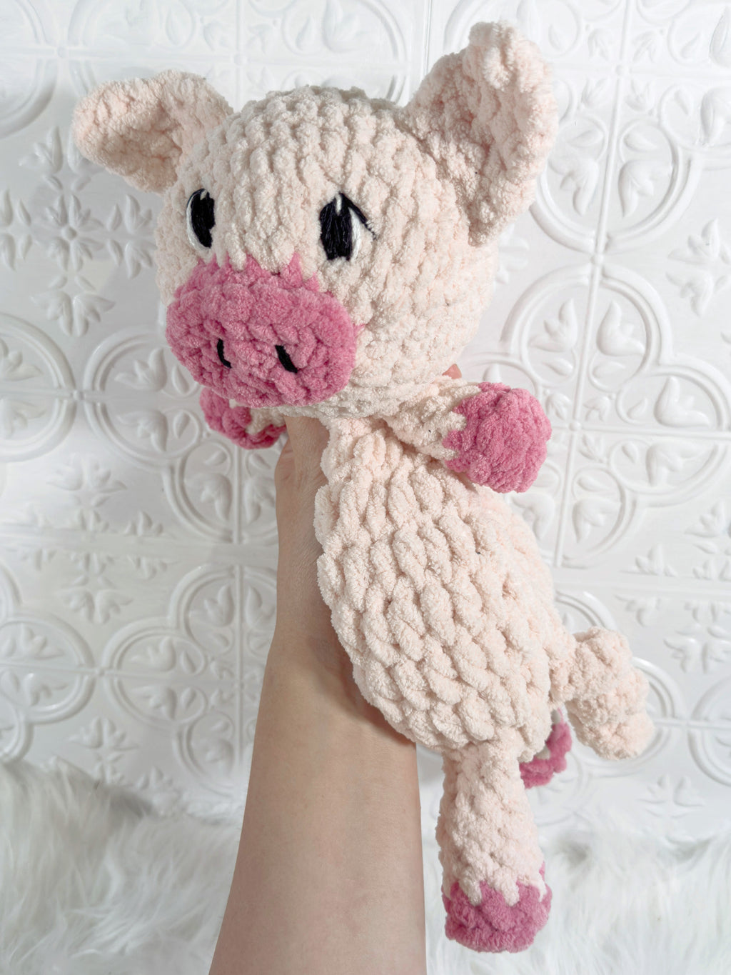 A handmade crocheted plush pig toy held up by a person's arm against a white embossed tile wall.