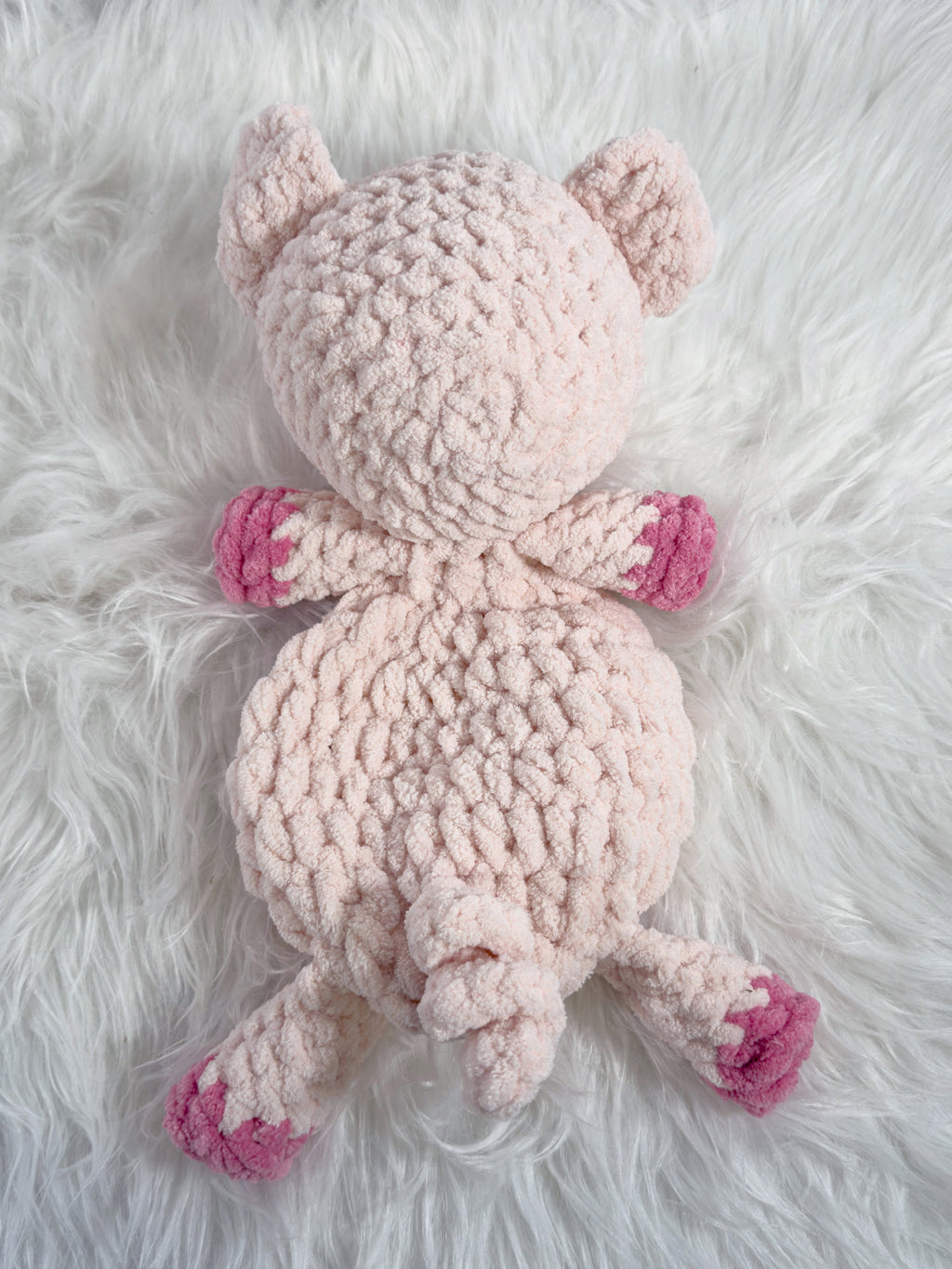 A light pink crocheted plush pig toy with darker pink paw pads, lying on a white faux fur background.