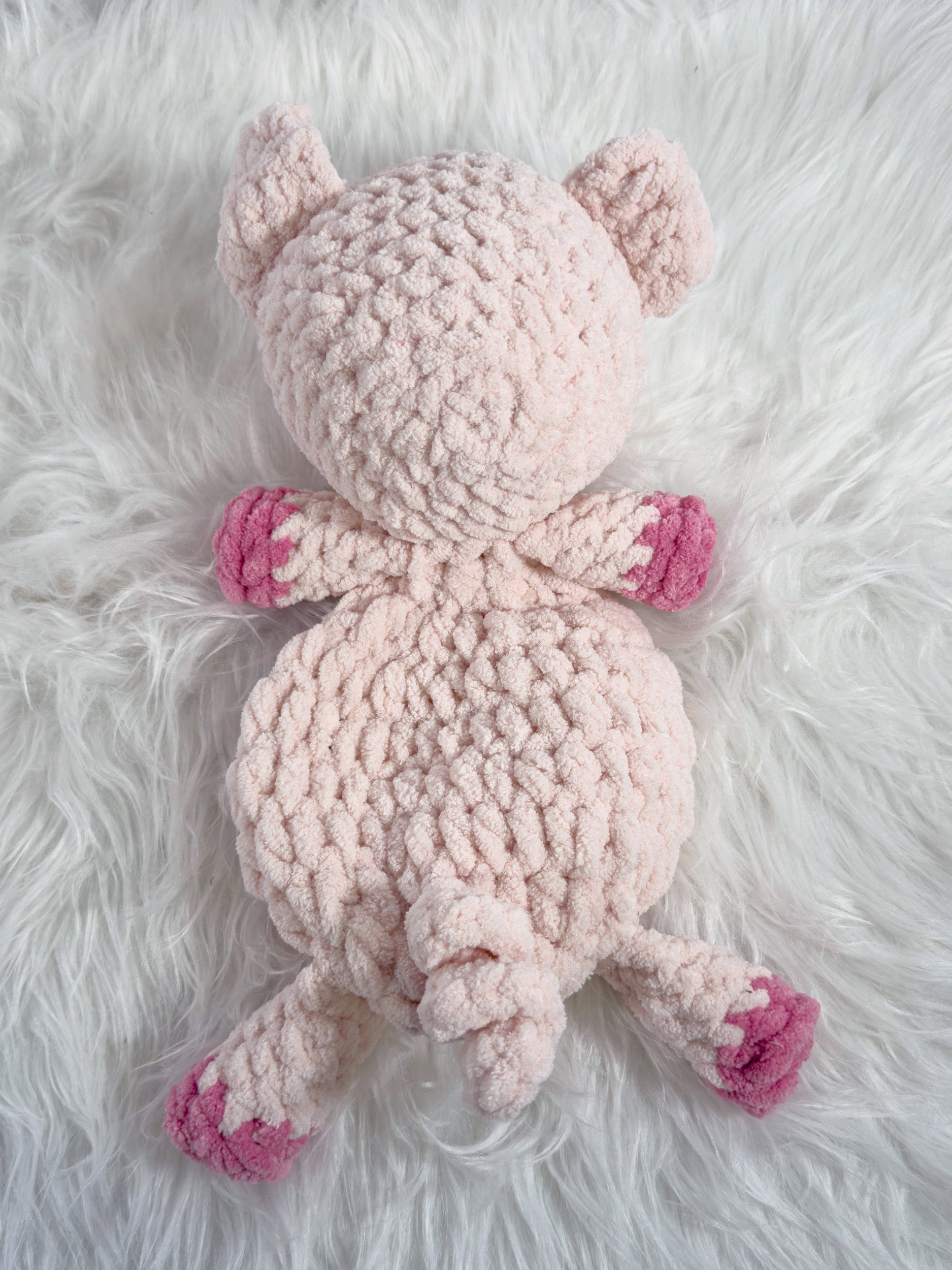 A light pink crocheted plush pig toy with darker pink paw pads, lying on a white faux fur background.