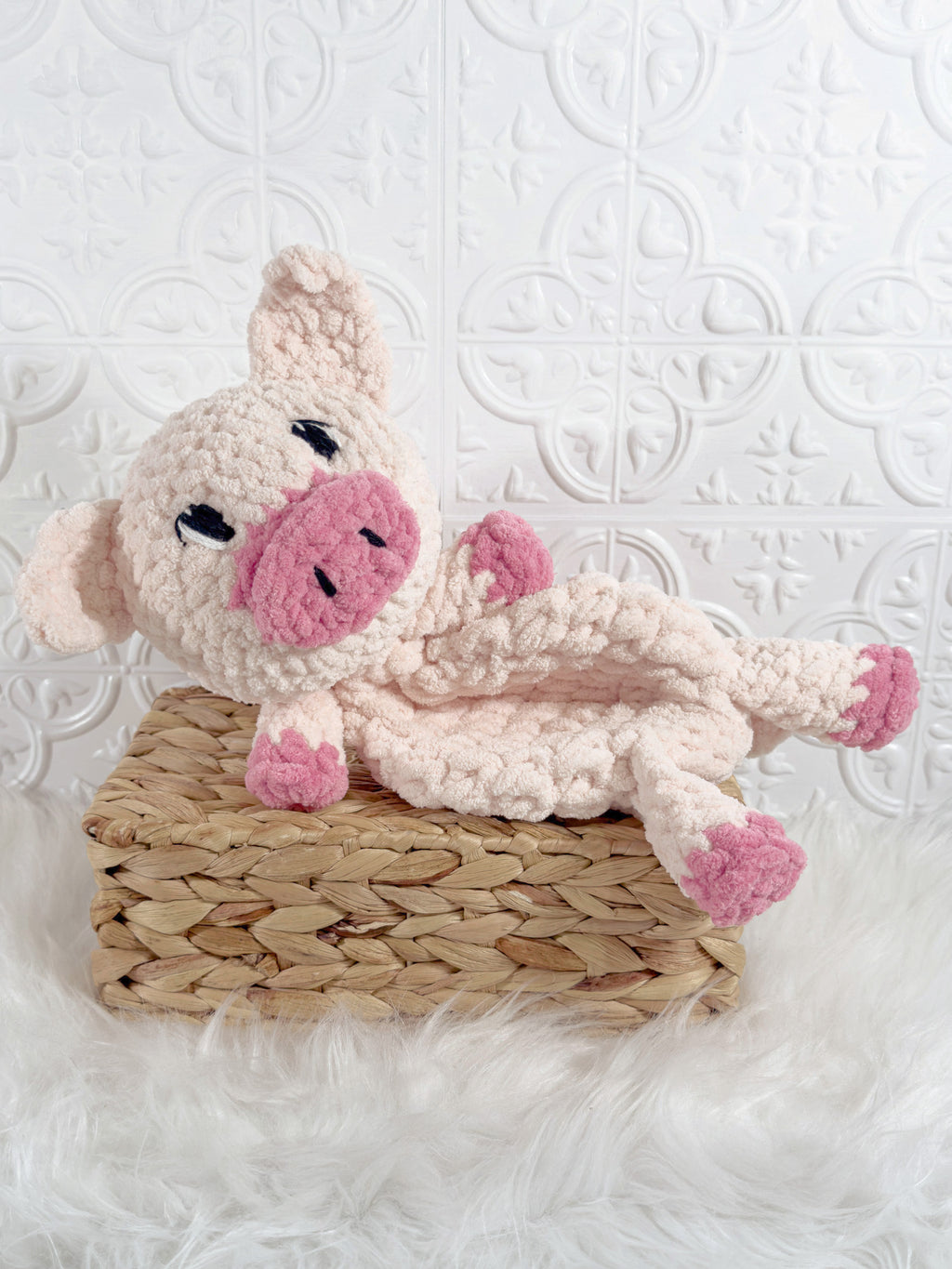 A crocheted plush pig toy with a pink snout and pink hooves, resting on a woven straw basket over a white faux-fur surface against a white patterned wall.